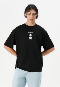 Schwarzes, übergroßes T-Shirt aus Baumwolle mit kurzen Ärmeln. Mit einem weißen "REPLAY"-Logo und grafischen Akzenten, die zentral auf der Brust platziert sind.
