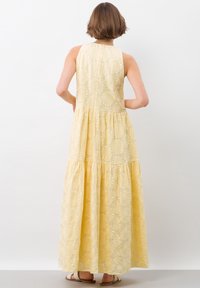 Une femme aux cheveux courts porte une robe maxi jaune à motifs floraux, montrant le dos et les volants du tissu sur un fond clair.