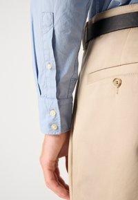 Camisa azul clara de botões, com botões nos punhos, combinada com calças beiges que possuem um bolso traseiro e um cinto preto, apresentando uma textura suave.