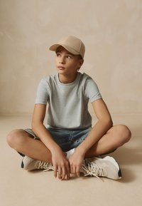 Maglietta grigio chiaro e pantaloncini di jeans blu indossati con un berretto beige. Scarpe da ginnastica beige e crema con texture. Posizione seduta con le gambe incrociate.