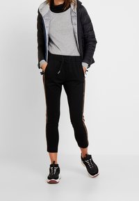 Sorte joggebukser med snoretræk i taljen, med gyldne sidest stripes og parret med en grå langærmet bluse og en sort pufferjakke.