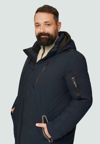 Marineblauer Parka mit Kapuze, mit Reißverschlusstaschen, schwarzen Akzenten und einer weichen Textur. Verfügt über ein Logo am Ärmel und kontrastierende Reißverschlussdetails.