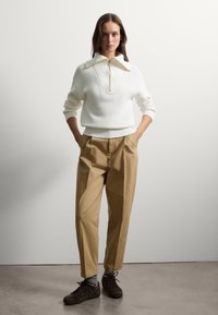 Witte ribgebreide trui met een half-zip kraag, gecombineerd met beige broeken met een relaxte pasvorm en geplooide voorkant, complementair met donkere schoenen.
