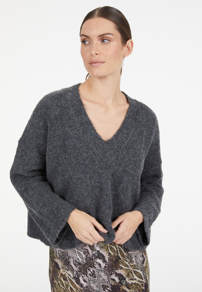 Grauer, fluffiger Strickpullover mit V-Ausschnitt, herunterfallenden Schultern und Glockenärmeln. Getragen über einem gemusterten, figurbetonten Rock.