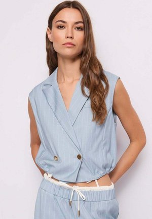 Donna con capelli lunghi e castani che indossa un blazer smanicato a righe sottili azzurro chiaro e pantaloni con coulisse abbinati, su uno sfondo neutro.