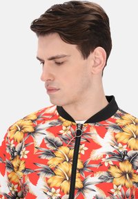 Giacca bomber floreale con uno sfondo rosso vibrante, motivi di ibisco giallo e nero, colletto a coste nere e chiusura con zip.