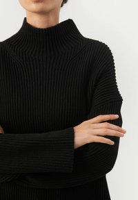 Schwarzer gerippter Pullover mit hohem Kragen, aus strukturiertem Stoff und breiten Bündchen, die Arme vor der Brust verschränkt, betont das einzigartige Design.