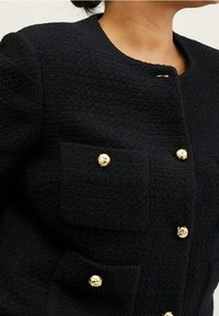 Kvinde iført en sort struktureret jakke med runde guldbuttons på frontlommerne og lukningen, som viser detaljer ved halsudskæringen og skuldrene.