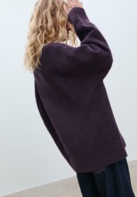 Übergroßer lila Pullover mit langen Ärmeln, gerippten Bündchen und weicher Textur. Kombiniert mit dunklen Jeans, vor einem hellen Hintergrund.