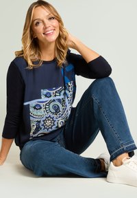 Marineblaues langärmeliges Oberteil mit einem bunten Paisley-Muster auf der Vorderseite, kombiniert mit blauen Jeans und weißen Turnschuhen. Glatte Stoffstruktur.