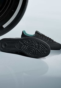 Baskets Adidas noires avec doublure intérieure turquoise, présentées avec la semelle vers l'avant, à côté d'un grand pneu noir sur un fond uni.