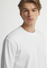 Weißes Langarmshirt aus Baumwolle mit Rundhalsausschnitt und entspannter Passform. Glatte Textur mit dezenten Stichdetails an den Ärmeln.
