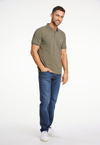 Polo shirt verde oliva con colletto e maniche corte, abbinato a jeans blu e sneakers bianche, su uno sfondo neutro.