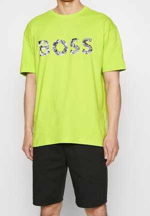 Neonově zelené bavlněné tričko s volným střihem, s velkým, maskáčovým logem "BOSS" na přední straně. Nosené s černými šortkami.