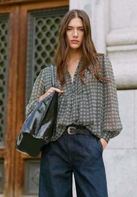 Borsa a spalla in pelle verniciata nera con borchie, abbinata a una blusa verde a motivi e jeans a gamba larga blu scuro, posizionata su uno sfondo strutturato.