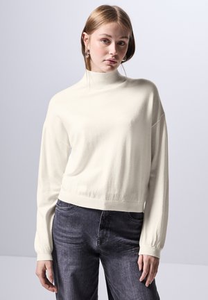 Gekürzter, langärmeliger Rollkragenpullover aus weichem, cremefarbenem Stoff. Mit geripptem Saum und Bündchen. Kombiniert mit dunkelgrauen, locker sitzenden Jeans.