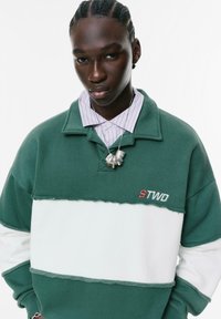 Felpa a blocchi di colore verde e bianco con colletto, caratterizzata da orli sfrangiati e logo ricamato 'STWD'. Indossata sopra una camicia a righe.