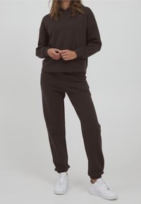 Donkerbruine sweatshirt met een capuchon en ribgebreide manchetten, gecombineerd met bijpassende joggers met elastische enkels, gedragen met witte sneakers.