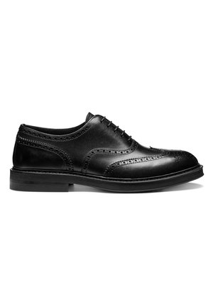 Zapato brogue de hombre de cuero negro con cierre cerrado, perforaciones decorativas y tacón apilado sobre fondo blanco.