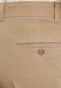 Pantaloni khaki con una texture liscia, dotati di una tasca posteriore con chiusura a bottone e dettagli cuciti lungo il cinturino e il bordo della tasca.
