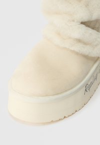 Bežīgas zamša zābaki ar pūkainu augšdaļu, biezā baltā zole un nelielu tekstūras atšķirību. Sānos ir "Replay" zīmols.