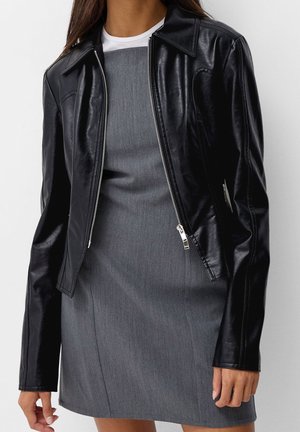 Femme portant une veste en cuir noire à fermeture éclair par-dessus une robe grise sans manches avec un haut blanc en dessous, debout devant un fond uni.