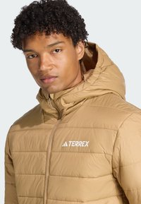 Ung mand med kort, krøllet hår iført en lysebrun, hættetrøje-jakke med TERREX-logoet. Neutral baggrund.
