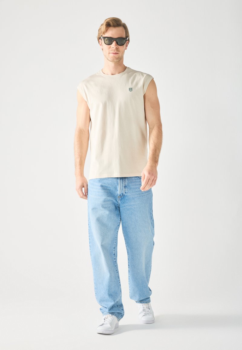 Beige sleeveless t-shirt with a green logo, paired with light-wash baggy jeans and white sneakers. Black sunglasses complete the look.