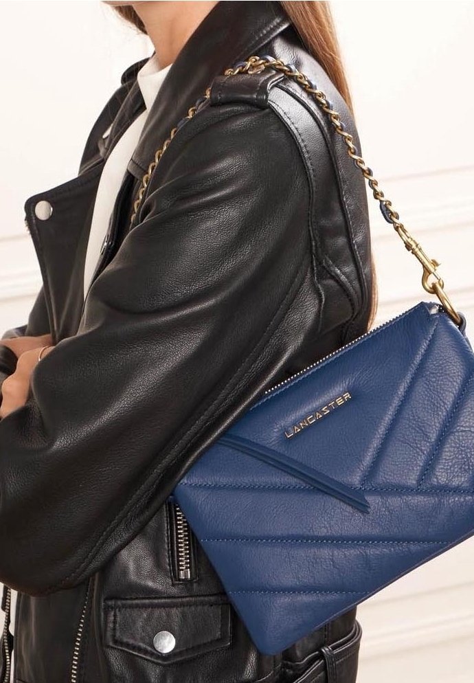 Femme portant une veste en cuir noire et un sac à main bleu Lancaster avec une bandoulière en chaîne dorée sur l'épaule.