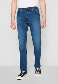 Jeans de mezclilla azul con un diseño de pierna recta, que presentan bolsillos frontales, un cierre de botón y un ligero desgaste. Llevados con zapatillas negras.