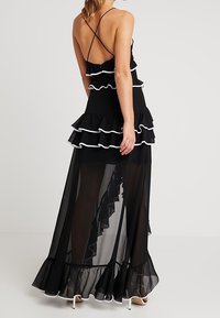 Schwarzes Maxikleid mit gestuften Rüschen, durchsichtiger Überlagerung und weißen Akzenten. Verfügt über verstellbare Spaghettiträger und ein tailliertes Oberteil.