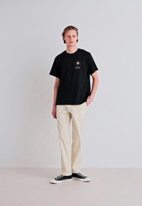 Tricou din bumbac negru cu grafică de soare, pantaloni crem și pantofi sport negri. Design simplu, croială lejeră, mâneci scurte și branding subtil.