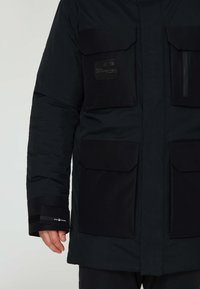 Svart isolerad jacka med flera stora fickor, en ficka med dragkedja på bröstet och kardborre-accenter på ärmarna. Tillverkad av slitstarkt, vattenavvisande material.