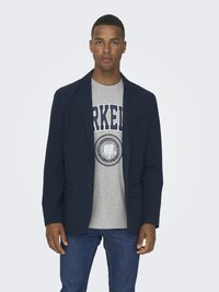 Marinblå blazer med två knappar och framfickor, bärs över en grå t-shirt med en cirkulär logotyp. Blå jeans fulländar outfiten.