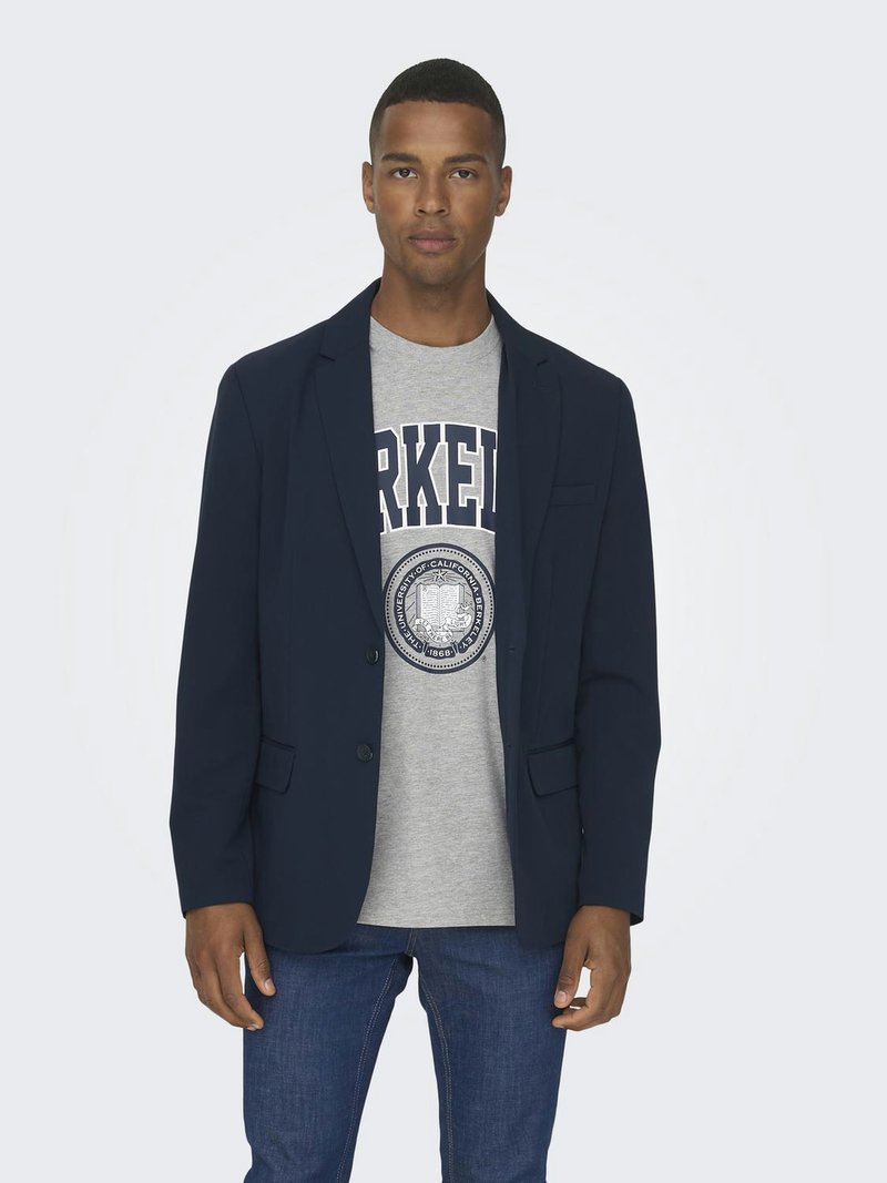 Blazer blu navy con due bottoni e tasche frontali indossato sopra una maglietta grigia con un logo circolare. Jeans blu completano l'outfit.