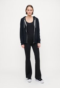 Svart hettejakke med hvite snorer, svart tank top, flagrende svarte bukser og svarte joggesko. Modellen står mot en enkel bakgrunn.