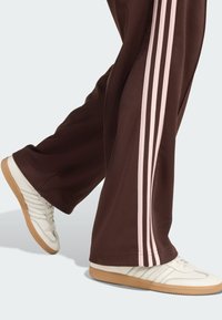 Brązowe spodnie dresowe z różowymi paskami po bokach, zestawione z białymi sneakersami z trzema jasno szarymi paskami i beżową gumową podeszwą.