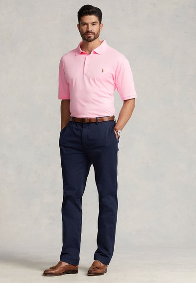 Rožinis polo marškinėlis trumpomis rankovėmis, tamsiai mėlynos kelnės ir rudi odiniai batai. Marškinėliuose yra mažas logotipas. Gerai prigludusi siluetas.