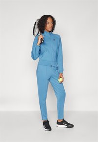 Helles blaues Sportanzug mit Reißverschlussjacke und Seitentaschen. Das Model hält einen Tennisschläger und einen Ball und trägt schwarze Turnschuhe.
