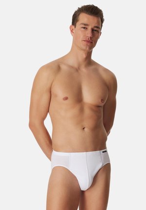 Young man standing against a white background wearing white brief-style underwear, arms positioned behind his back, neutral expression.