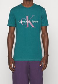 Tyrkysové bavlněné tričko s velkým růžovým a bílým logem "Calvin Klein Jeans", kulatým výstřihem a krátkým rukávem. Hladký povrch, volný střih.
