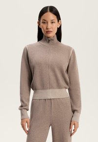 Tovarna kapucna majica v taupe barvi z visokim ovratnikom, rebrastimi manšetami in rebrastim robom. Mehak značaj s kontrastnimi rebrastimi poudarki vzdolž ramen.