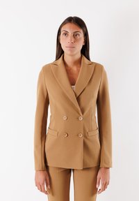 Blazer marrone doppiopetto con rever a punta, tasche frontali e tessuto morbido. Abbinato a pantaloni coordinati per un look coordinato.