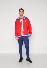Red Nike jacket with blue accents, white Athletic shirt featuring red stripes, navy pants with yellow Nike logo, and black shoes.
