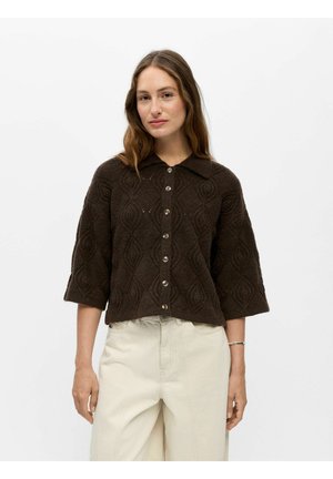 Femme portant un cardigan boutonné en maille texturée marron foncé avec des manches trois-quarts et un pantalon taille haute écru.