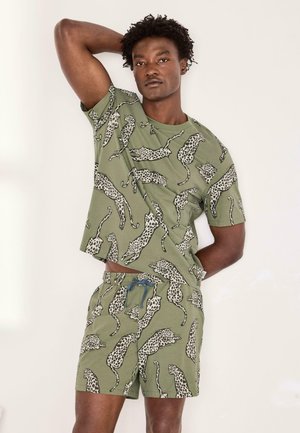 Mężczyzna ubrany w dopasowany oliwkowozielony T-shirt i szorty z czarno-białym leopardzim wzorem, pozujący z jedną ręką uniesioną za głową.
