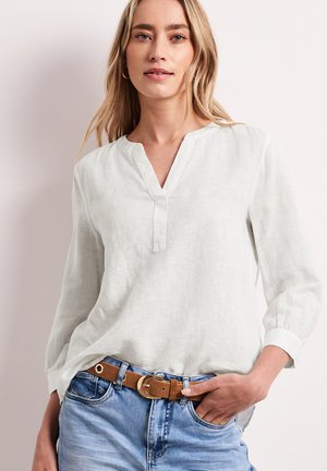 Blonde vrouw die een witte blouse met lange mouwen en V-hals draagt, met lichtblauwe spijkerbroek, gecombineerd met een bruine riem en gouden creolen.
