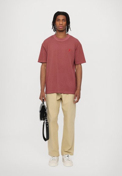 Camiseta corta de color marsala con cuello redondo y un pequeño logo rojo. Combinada con pantalones beiges y zapatillas blancas. Sosteniendo un bolso negro.