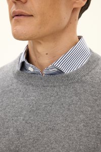 Grauer Pullover aus weichem Stoff mit einem Rippkragen, der über einem blau-weiß gestreiften Kragenhemd mit Knopfdetails getragen wird.