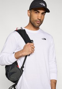 Μαύρο καπέλο και λευκό πουκάμισο με μακριά μανίκια, που φοριέται από ένα άτομο που κρατά ένα μαύρο και κόκκινο σακίδιο με ρυθμιζόμενους ιμάντες.